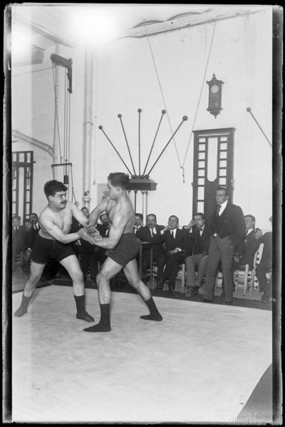 Match entre V. Huesta I Santasusagna en un assalt del campionat de lluita grecoromana de l'Ateneu Enciclopèdic Popular amb el jurat al fons — Co i de Triola, Josep Maria, 1884-1965